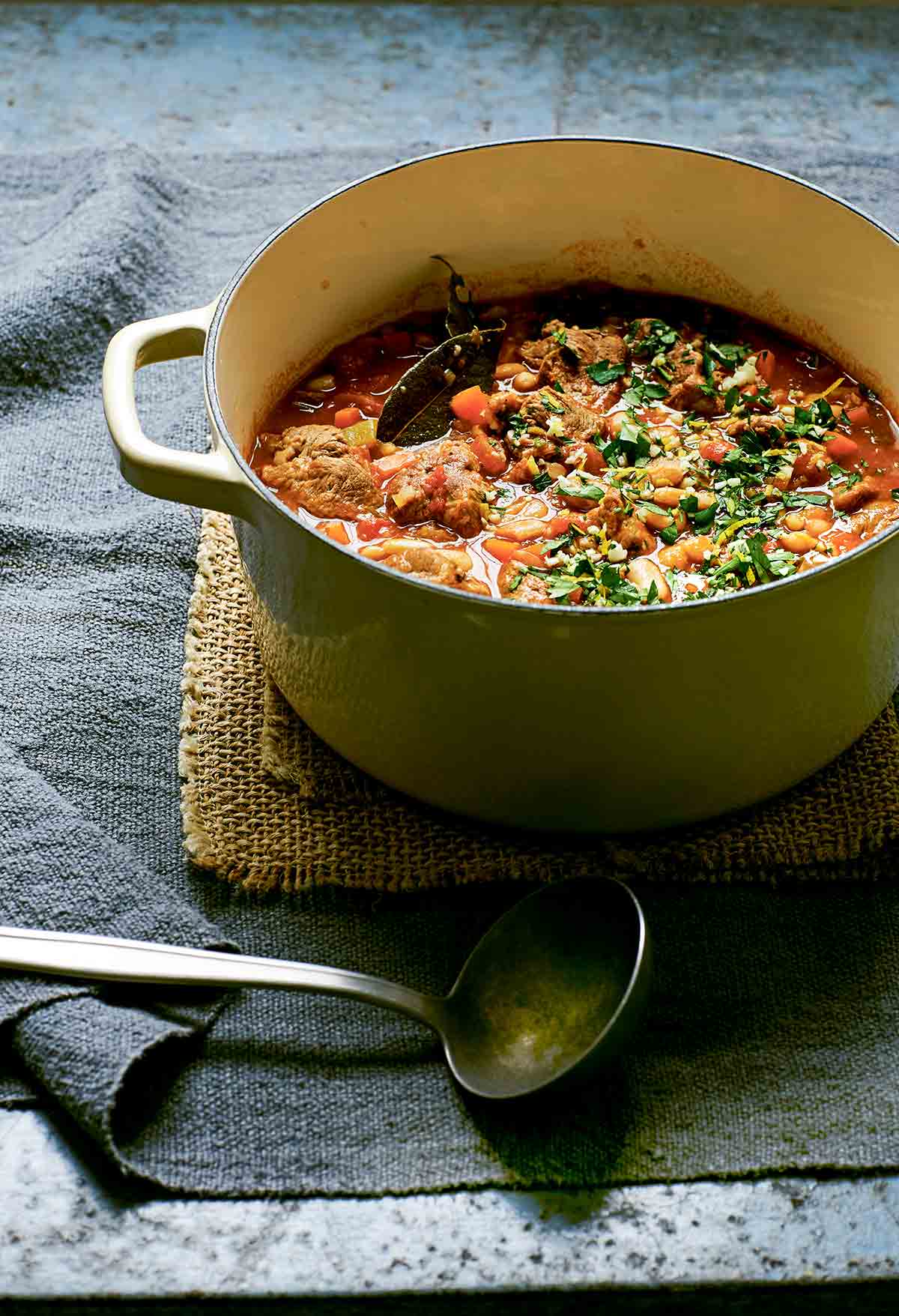 A round green Dutch oven filled with slow cooked lamb and cannellini beans on a brown burlap cloth.