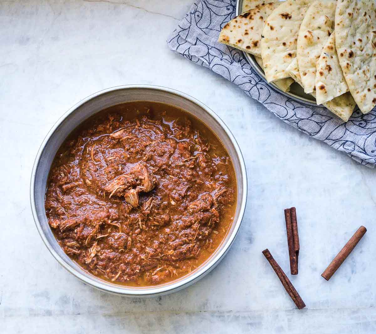 A bowl of slow cooker Indian-style chicken with cinnamon sticks and pieces of naan on the side.