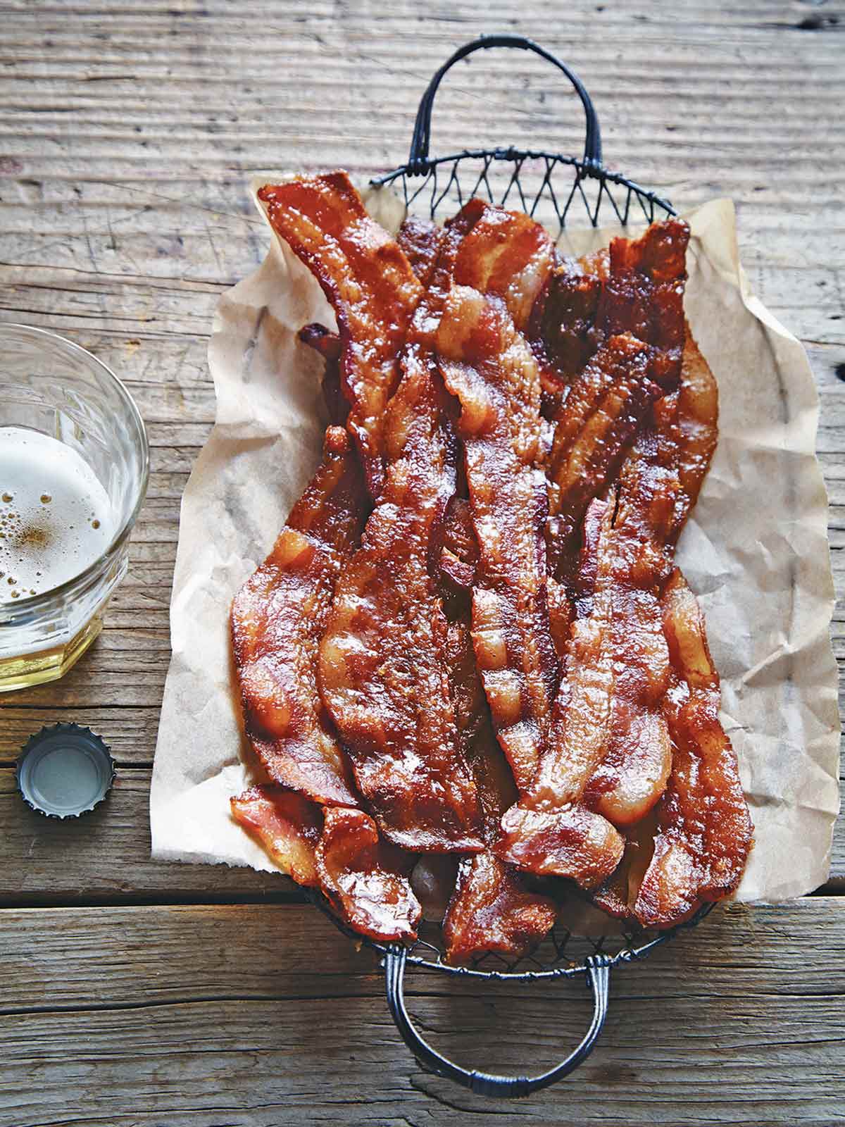 A wire basket lined with parchment and filled with sweet and spicy bacon with a glass of beer on the side.